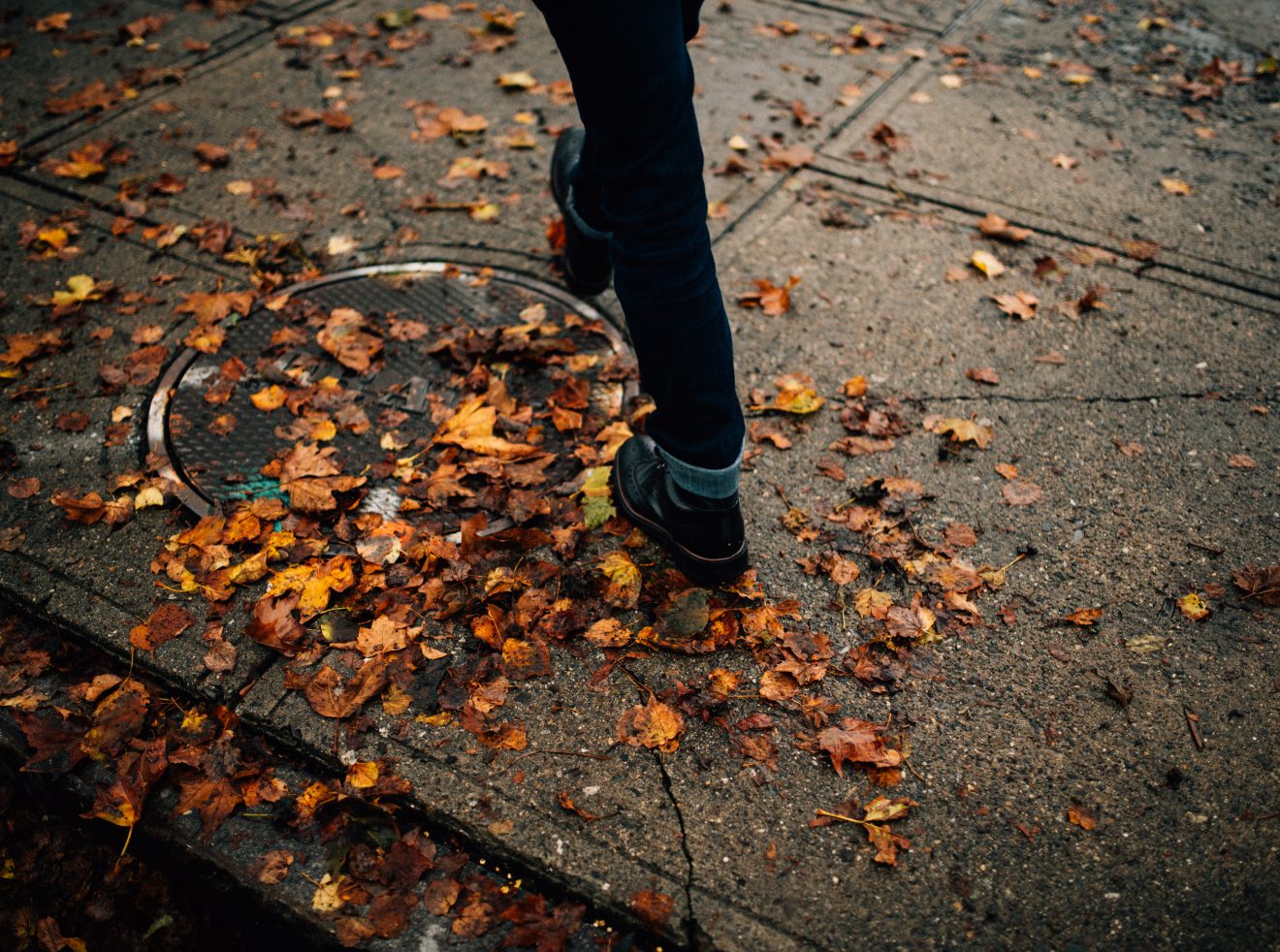 Man in boots walks on Salem sidewalk, stepping on fallen orange leaves.