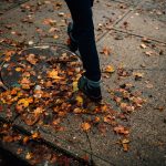 Man in boots walks on Salem sidewalk, stepping on fallen orange leaves.