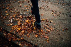 Man in boots walks on Salem sidewalk, stepping on fallen orange leaves.