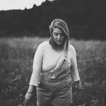 Girl in overalls walks through field, feeling the grasses and flowers.